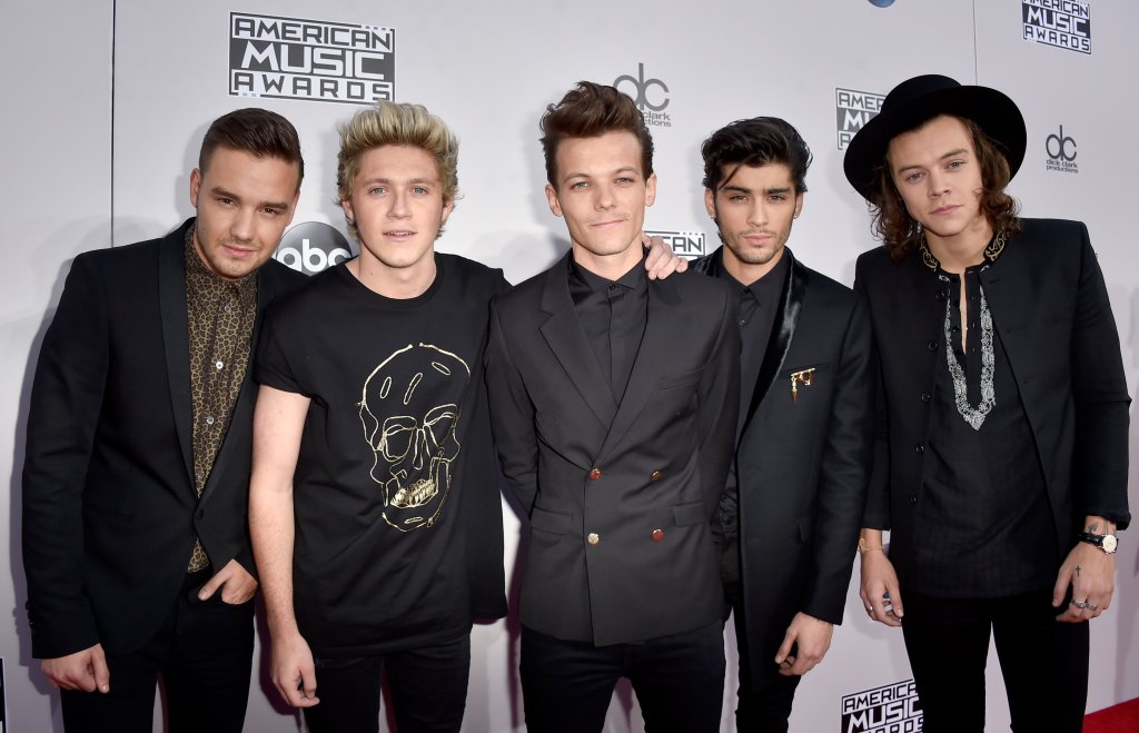 Foto dos integrantes do One Direction no tapete vermelho posando. Todos estão de preto e Harry está com um chapéu.
