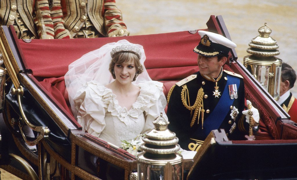 Princesa Diana e príncipe Charles em seu casamento em 1981, em carruagem vermelha, Charles com suas vestes de principe, e Diana com seu iconico vestido de noiva branco, com véu, babados, decote redondo. Os dois aparecem sorridentes.
