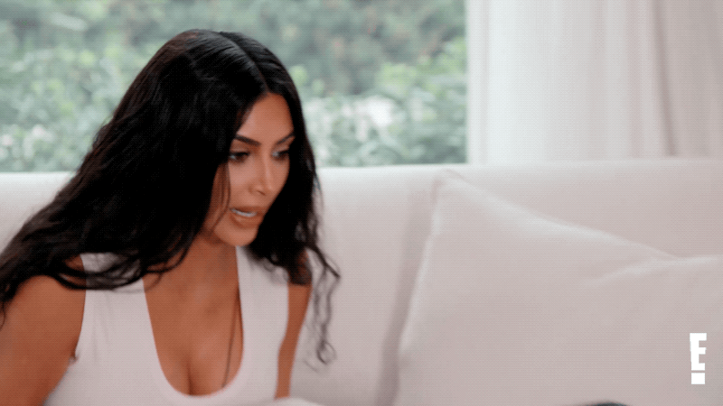 Kim Kardashian com blusa branca com decote fazendo expressão de surpresa e levando uma das mãos a boca. Ela esta sentada em um sofá branco.