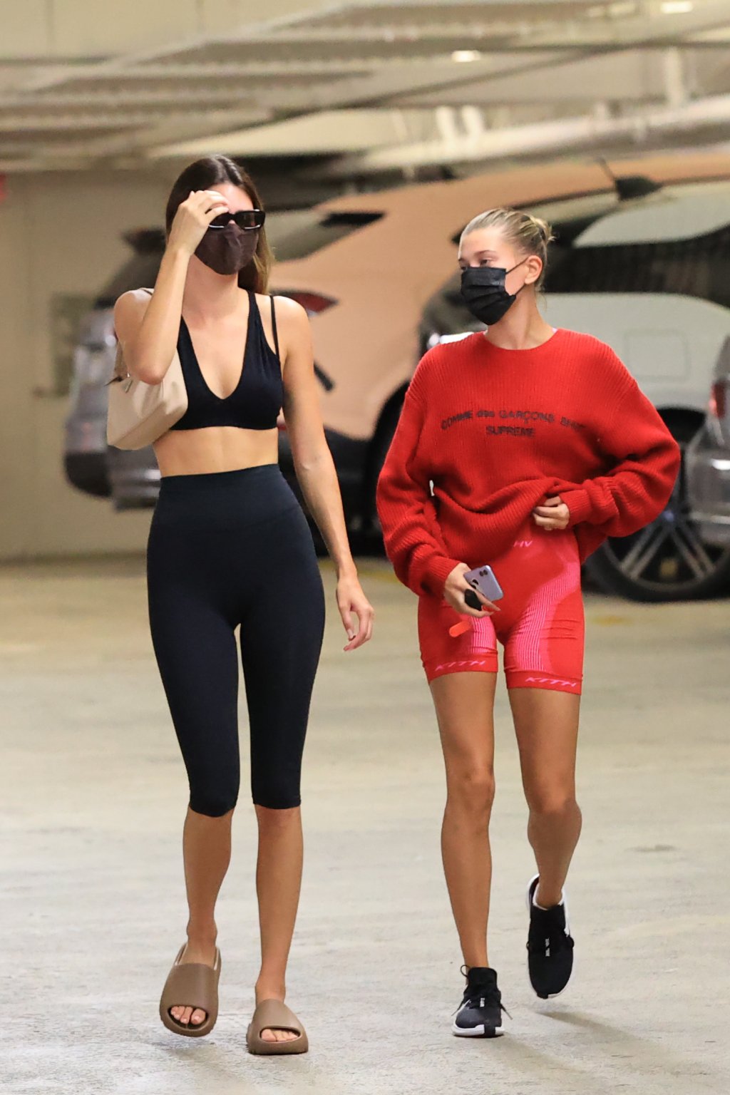 Kendall Jenner usando look preto formado por top de academia e biker shorts com chinelo slide e shoulder bag, e Hailey Bieber usando look vermelho formado por moletom, biker shorts e tênis