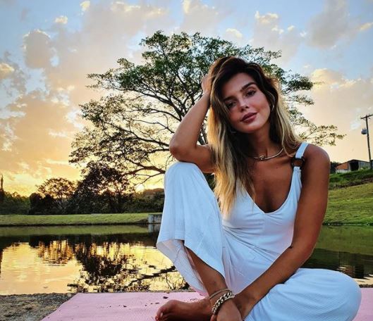 Giovanna Lancelotti posando para foto em uma paisagem natural com lago e pôr-do-sol de fundo com uma árvore e grama; ela está sentada uma roupa branca, o cotovelo apoiado no joelho e cabeça apoiada na mão