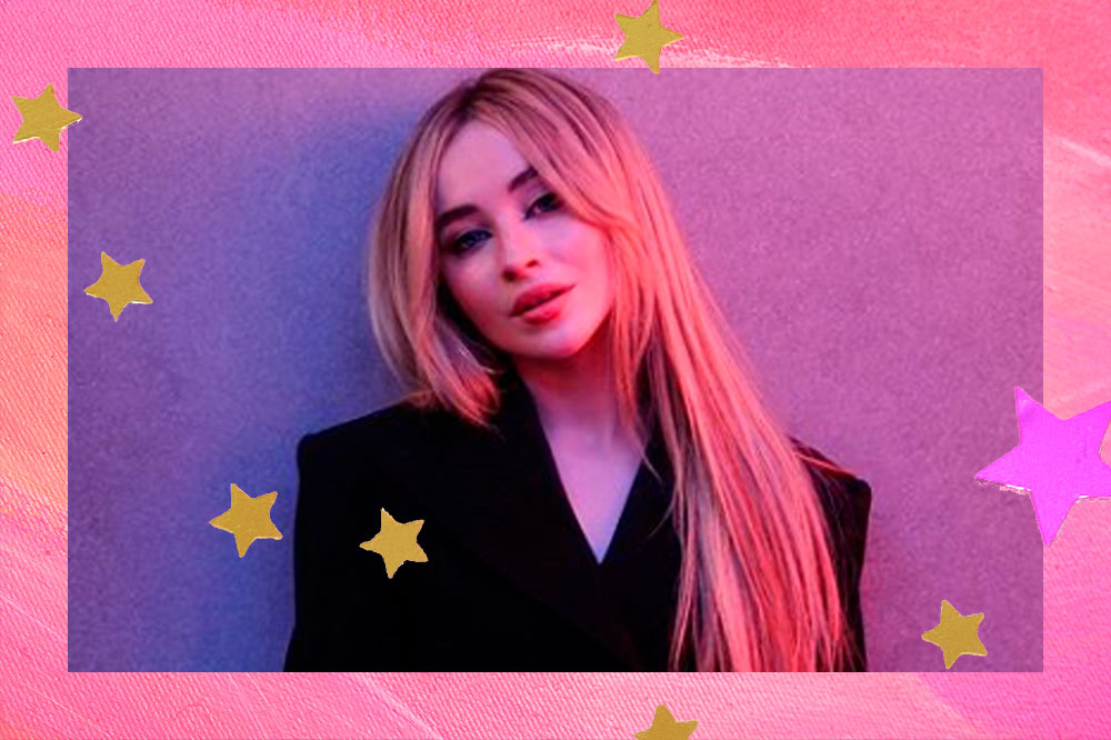 Sabrina está com cabelos longos e loiros, de roupa preta em uma parede om luzes roxas e rosas.