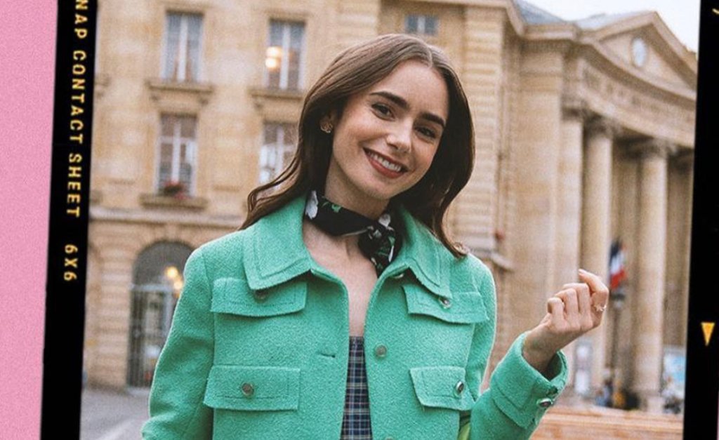 Lily Collins caracterizada como Emily; ela posa para foto sorrindo, com um lenço no pescoço, casaco verde e roupa xadrez por baixo