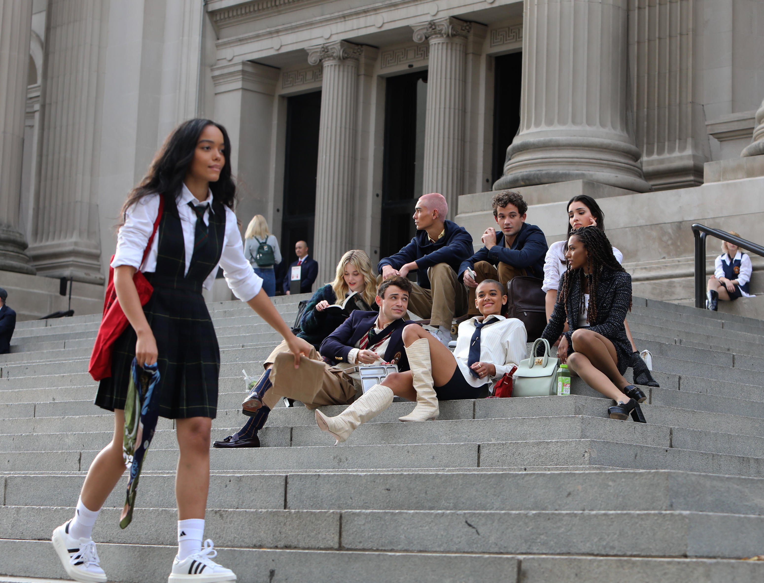 Elenco do reboot de Gossip Girl sentados na escadaria do MET, em Nova Iorque. Alguns atores estão olhando para tela ou sorrindo enquanto outros estão com expressões sérias. Uma da protagonista está passando por eles no primeiro plano da imagem
