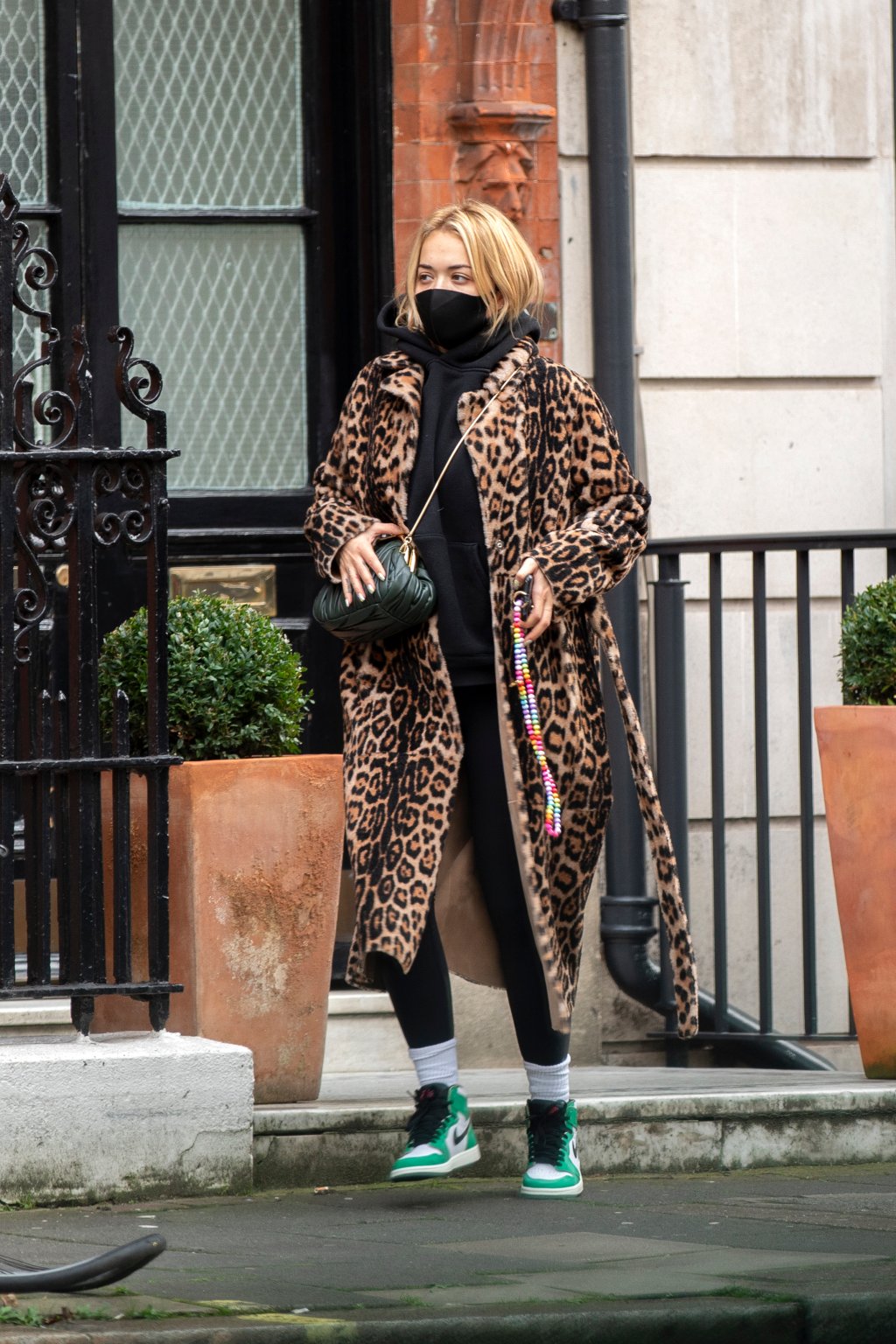 Rita Ora usando look com moletom preto, calça legging preta, tênis verde e branco de cano alto, sobretudo com estampa de oncinha e bolsa preta atravessada no ombro. Ela está com máscara de proteção preta, olhando para o lado, em uma pose espontânea.