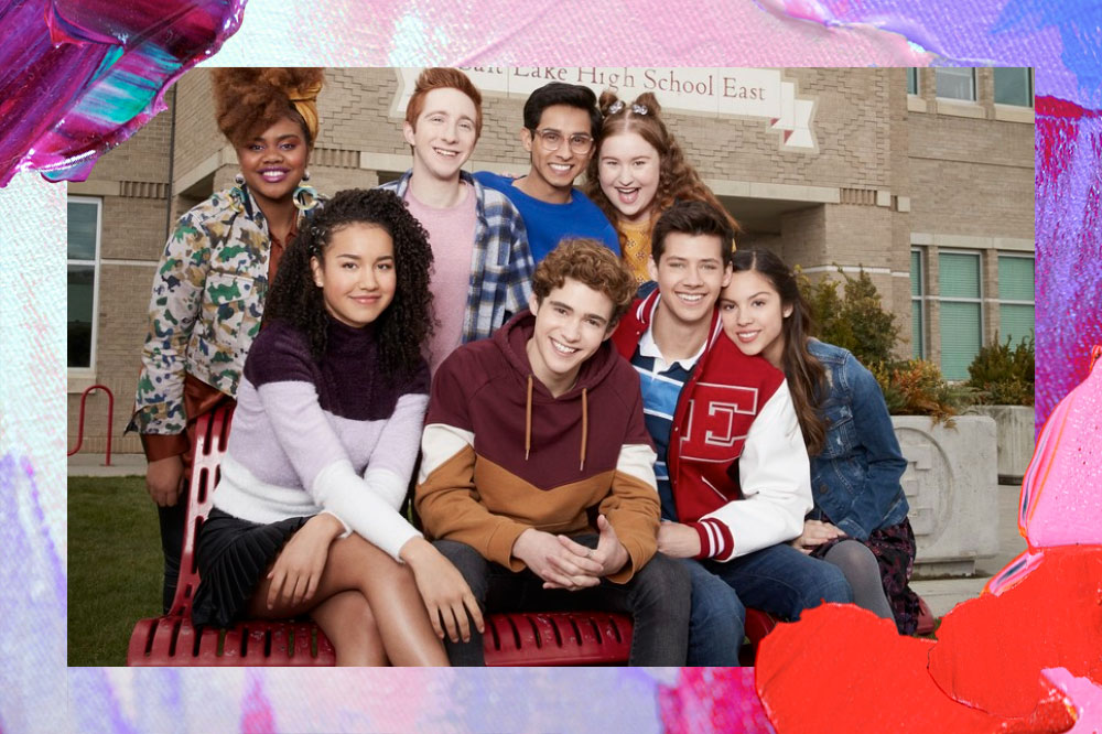 Elenco de High School Musical: The Musical: The Series juntos posando para foto; todos estão sorrindo e encostados um dos outros enquanto sentam em uma área aberta; a margem é uma textura de tintas nas cores rosa, roxo, azul, vermelho e branco