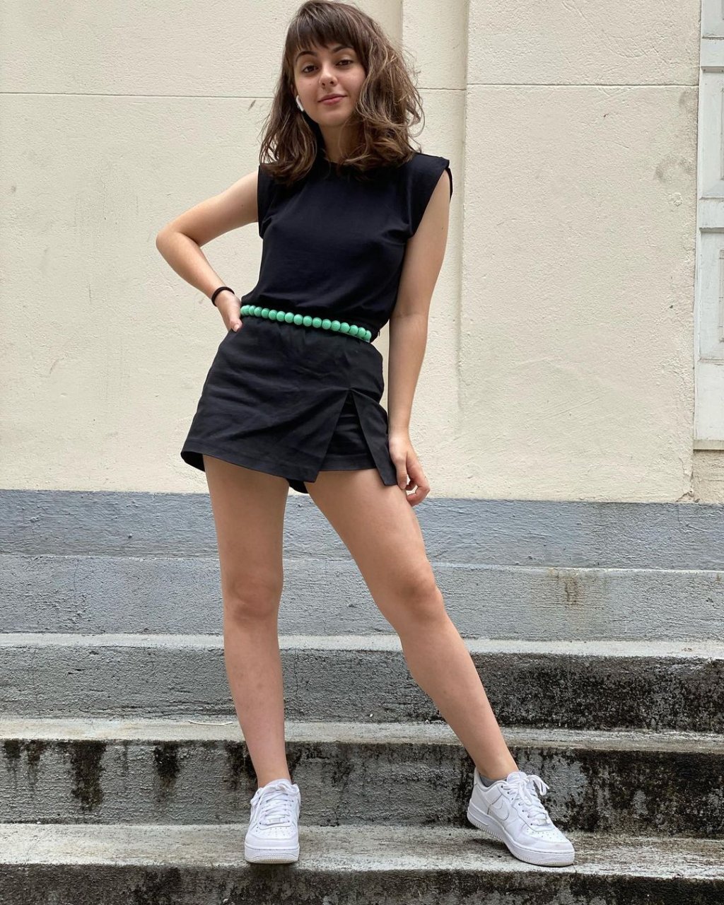 Klara Castanho usando muscle tee preta com short saia preto, tênis branco e cinto verde
