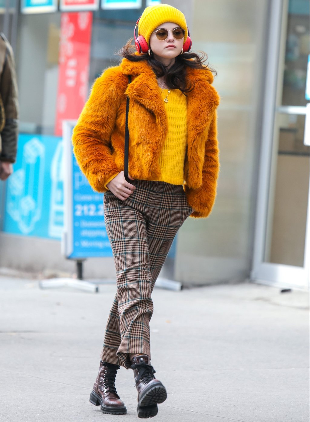 Selena Gomez andando na rua com gorro e suéter amarelos, casaco de pelos laranja, calça xadrez e coturno; ela está com a expressão séria e usa headphones vermelhos