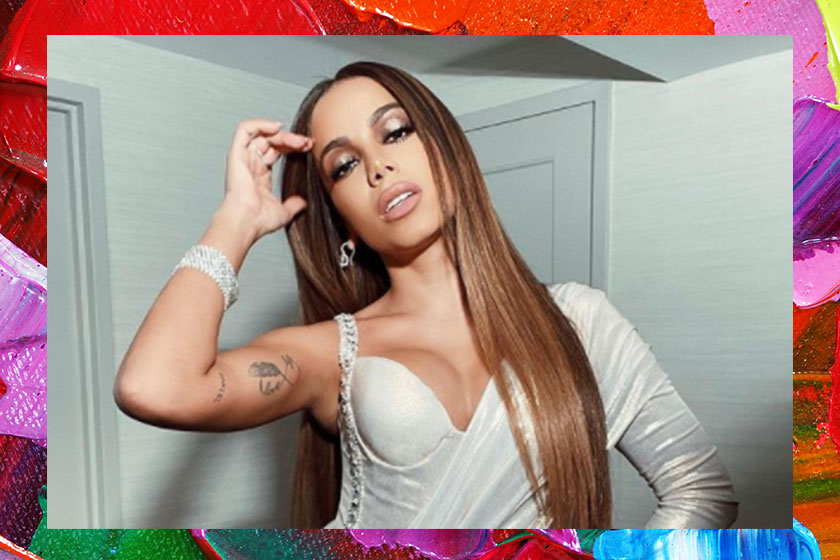 Anitta posando para foto com uma mão na cabeça e a outra na cintura; ela usa uma roupa branca com brilhos prateados