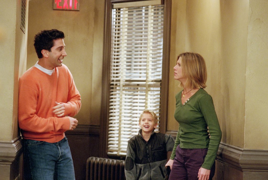 cena de Friends mostra, de um lado, Ross encostado em uma parede, com as mãos na barriga e sorrinho. De outro, Rachel olha para ele com cara de desaprovação. Ben está entre os dois, sorrindo