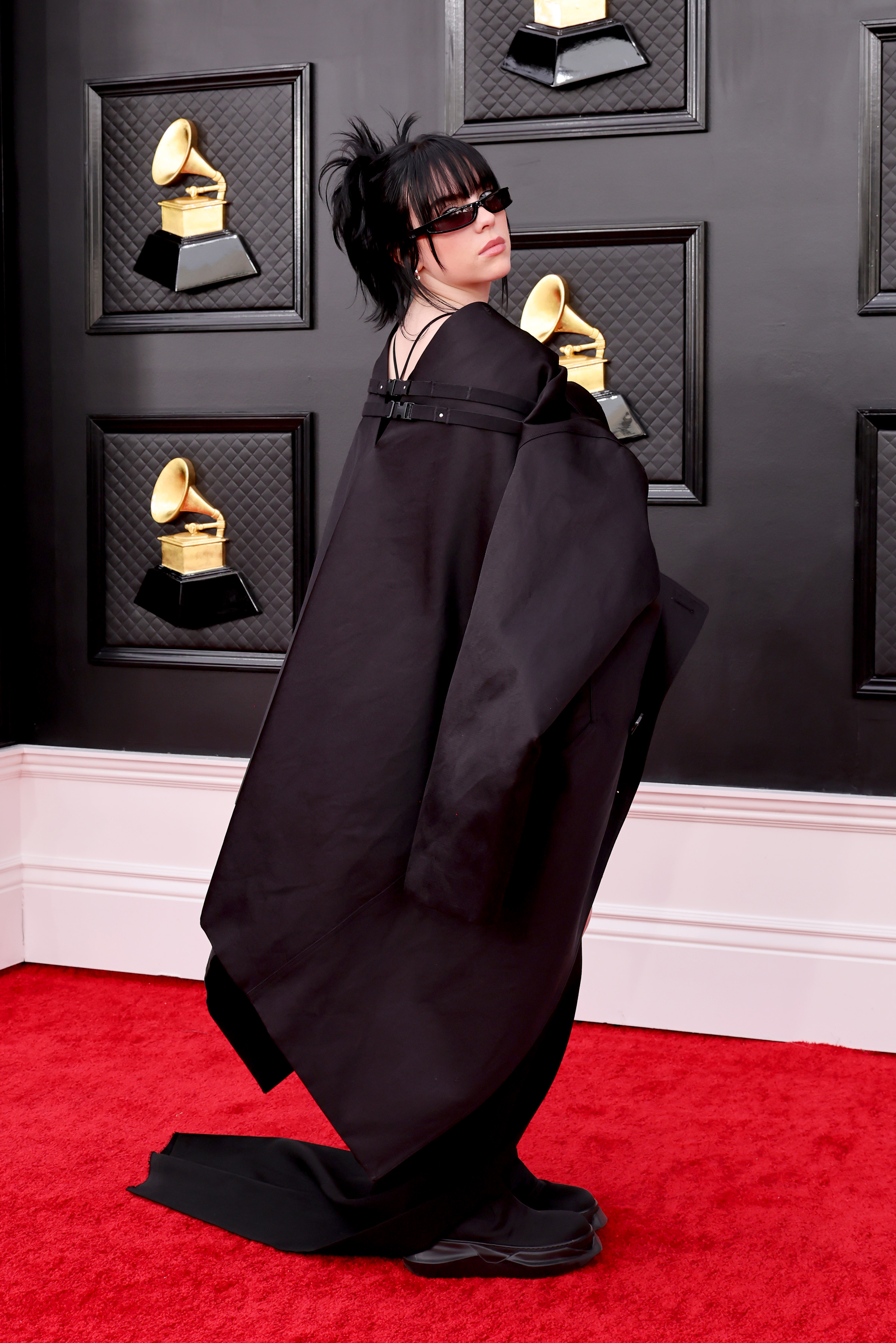 Imagem mostra Billie Eilish no tapete vermelho da 64ª edição do Grammy Awards usando um roupa-casulo preta. Ela está posando quase que costa e usa óculos escuro.