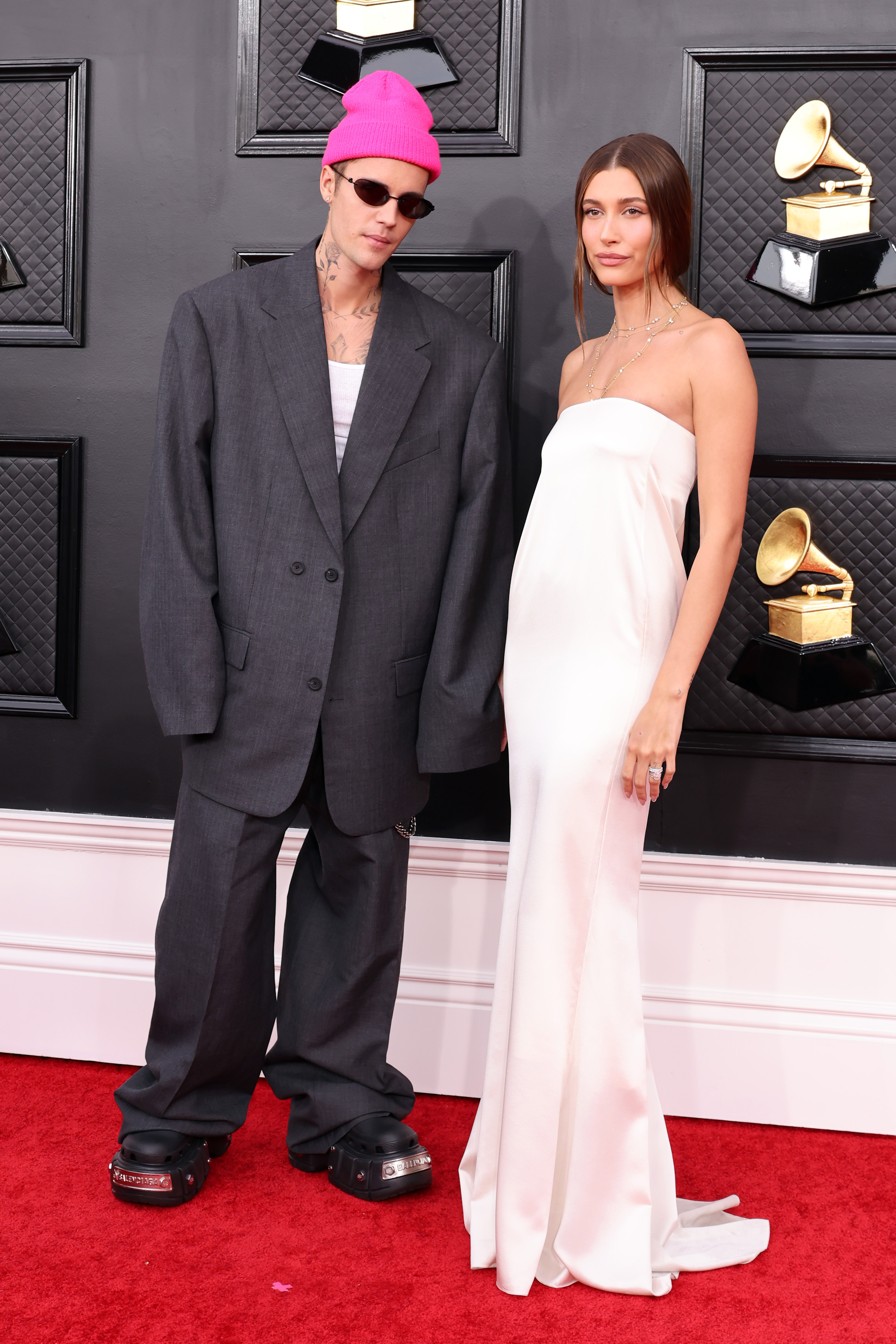 Imagem mostra Justin Bieber e Hailey Bieber posando juntos no tapete vermelho da 64ª edição do Grammy Awards. Justin usa óculos escuro, gorro rosa pink e um blazer e calça cinza escura com modelagem ampla e oversized. Enquanto Hailey um vestido branco de cetim sem alças com modelagem mais reta. Ambos olham para a câmera com expressão leve.