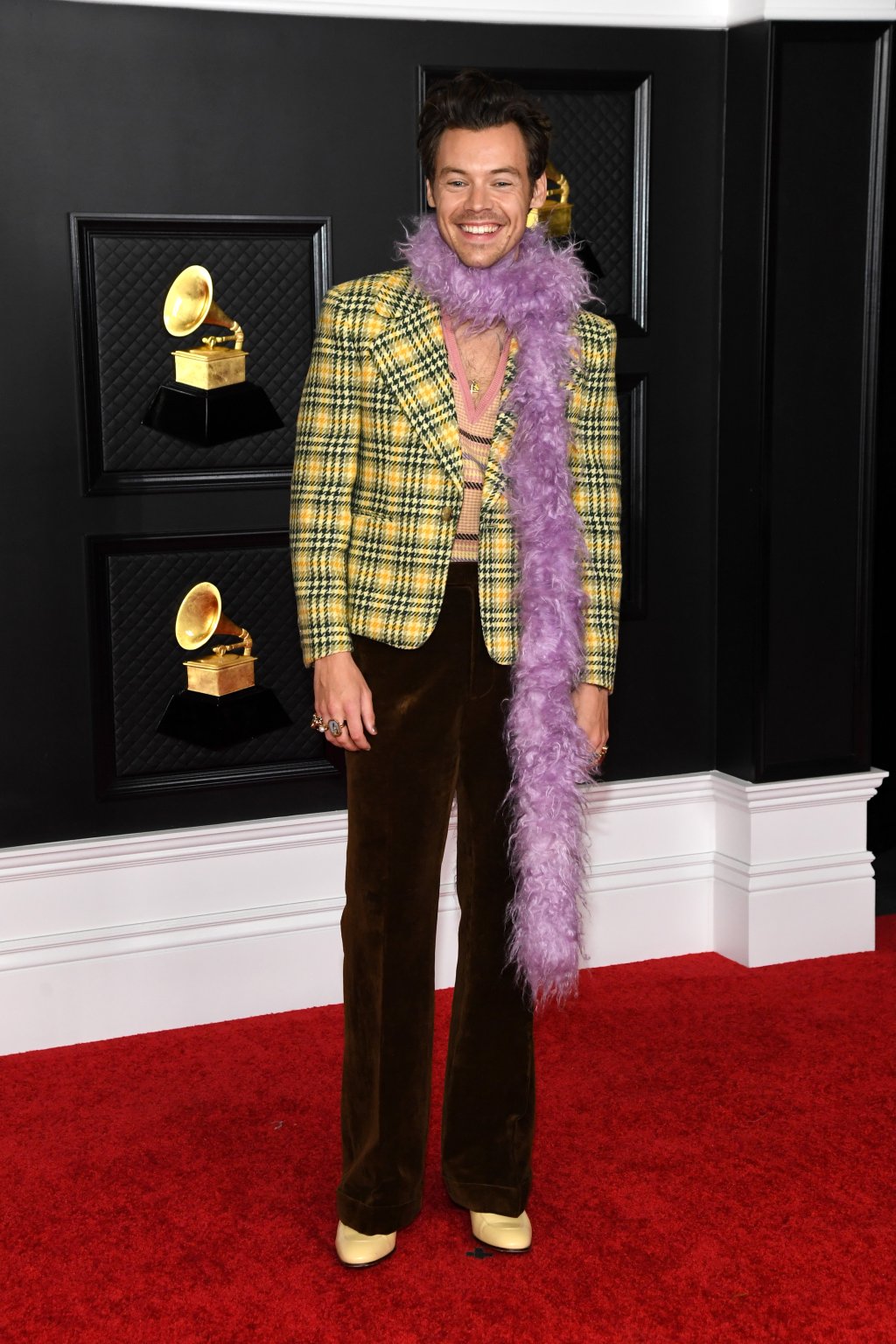 Harry Styles em pé, no tapete vermelho do Grammy 2021. Ele está usando um blazer xadrez amarelo e verde por cima de um colete de tricô, uma calça de veludo marrom, um sapato bege e um cachecol lilás de plumas. Ele está sorrindo e olhando para a câmera.