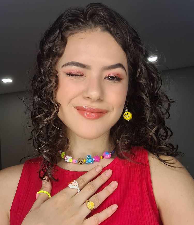 Maisa Silva com acessórios divertidos, com miçangas coloridas, posando para selfie piscando um dos olhos, usando regata vermelha com uma das mãos tocando o coração.