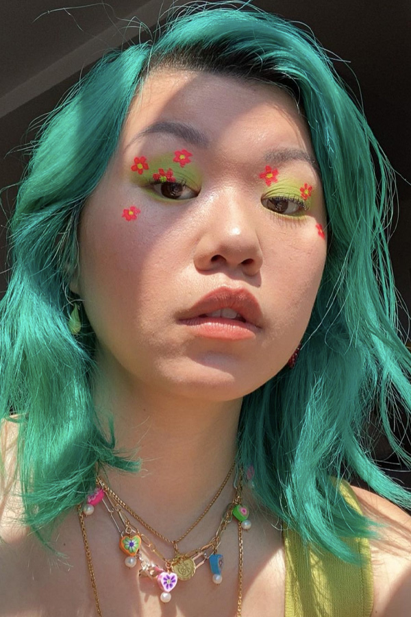 Inspiração de maquiagem colorida com flores de cor laranja e sombra esfumada verde, jovem com cabelo verde água posando para foto com close em seu rosto.