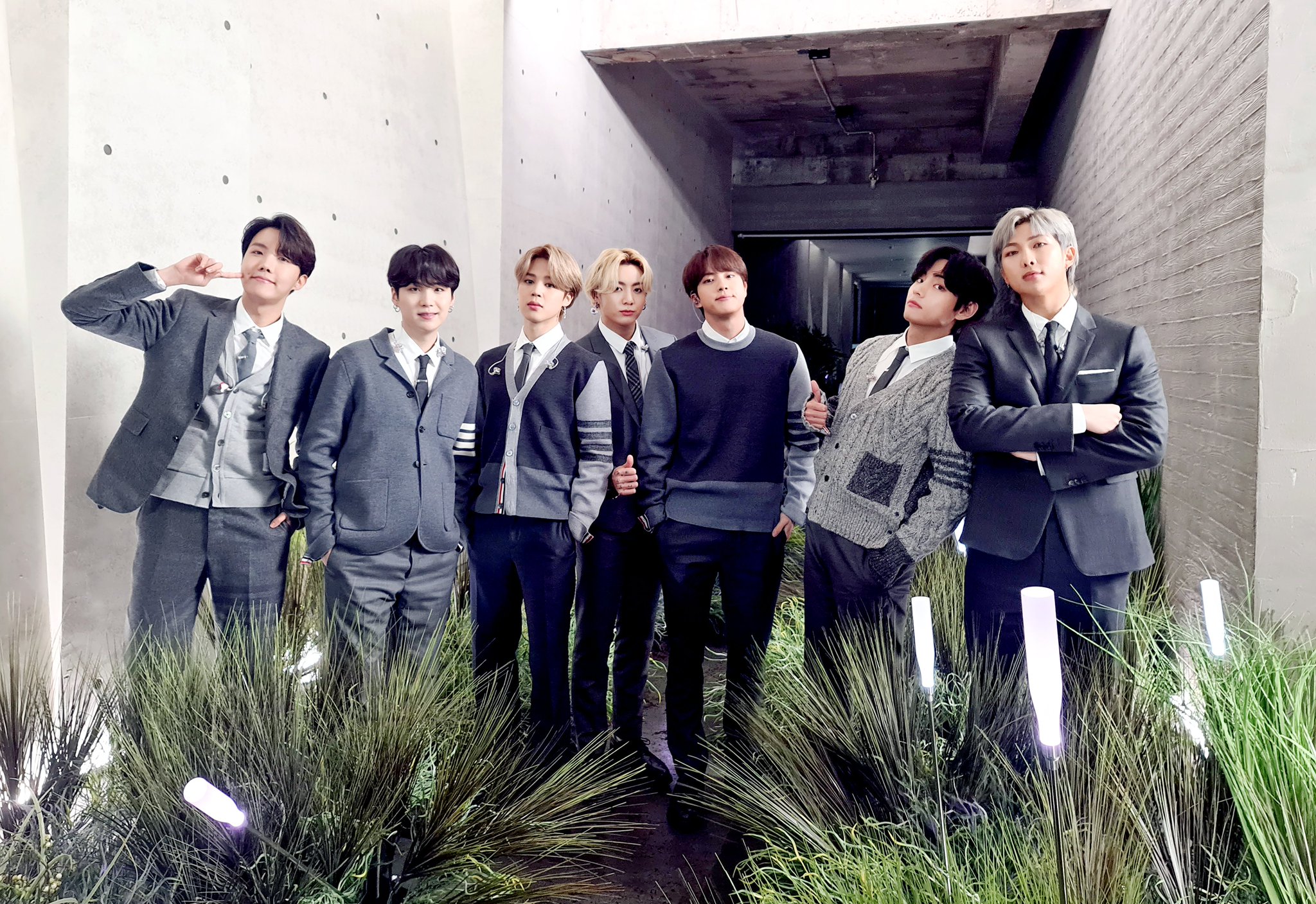 Grupo BTS posando para foto em meio a grama em roupas com tom de cinza e preto