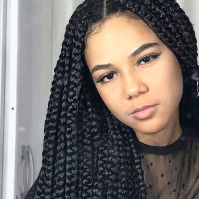 Box braids Jovem com a cabeça levemente inclinada para o lado esquerdo, com expressão séria e usando blusa preta transparente.