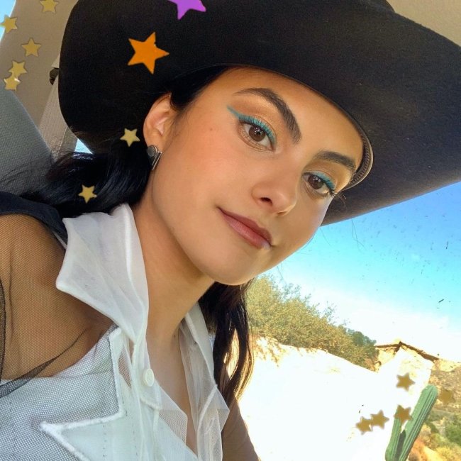 Atriz Camila Mendes com delineado verde, usando chapéu de cowboy preto, camisa branca com babado e posando para selfie com uma expressão tranquila