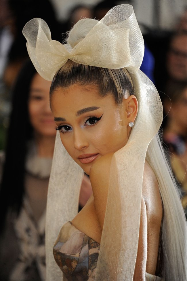 Ariana Grande posando para foto virada de lado, com expressão séria, com a cabeça levemente abaixada, com grande laço na cabeça e vestido cinza tomara que caia.