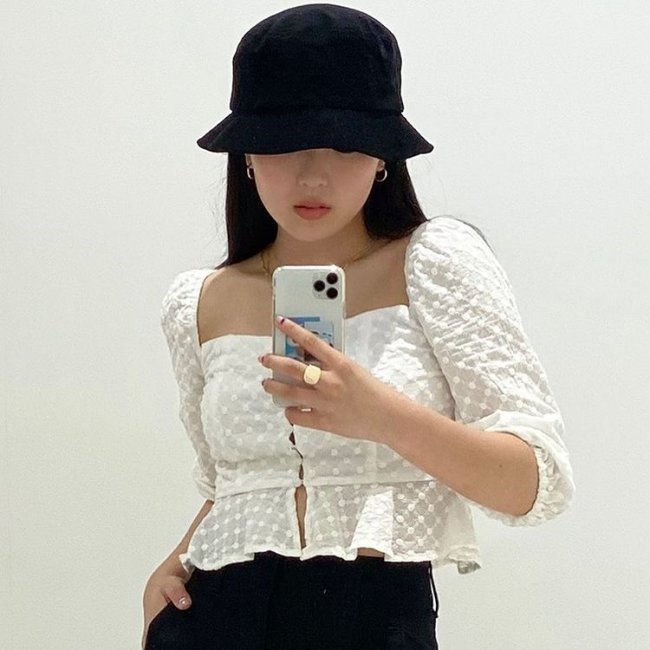Hina com Bucket Hat Hina, do grupo Now United, com Bucket Hat preto, usando blusa com manga bufante branca, foto da cintura para cima, mostrando o celular e com uma das mãos no bolso.