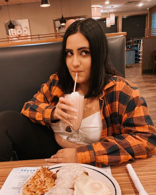 Jovem tomando milkshake usando camisa xadrez laranja e preta dentro de uma lanchonete, sentada em uma das mesas em um banco de couro preto posando para foto.