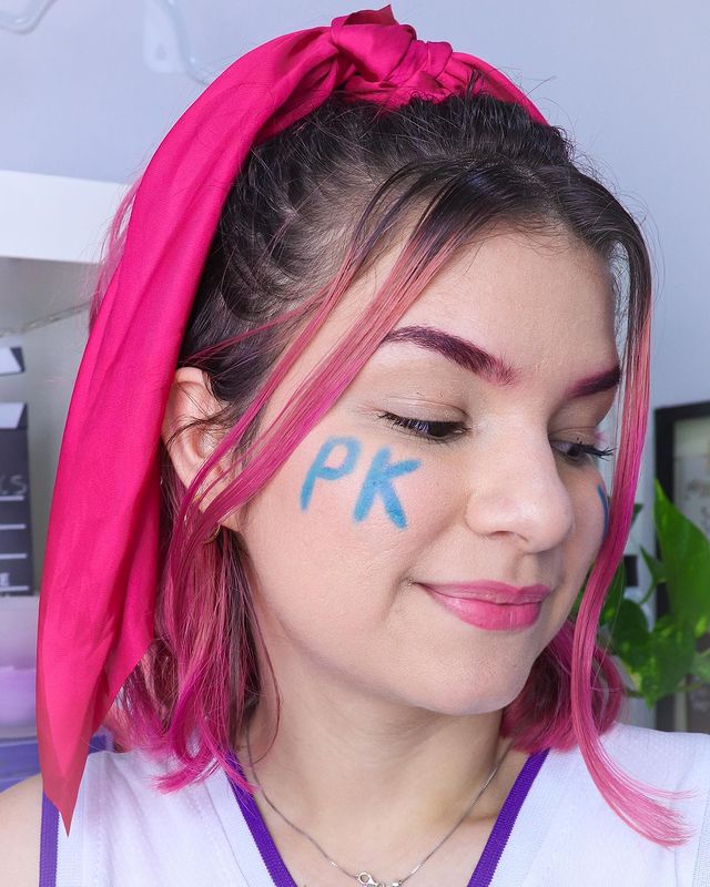Jovem olhando para baixo com as letras PK pintadas em sua bochecha de com tinta azul, os cabelo é rosa pink e está meio preso com um lenço rosa.