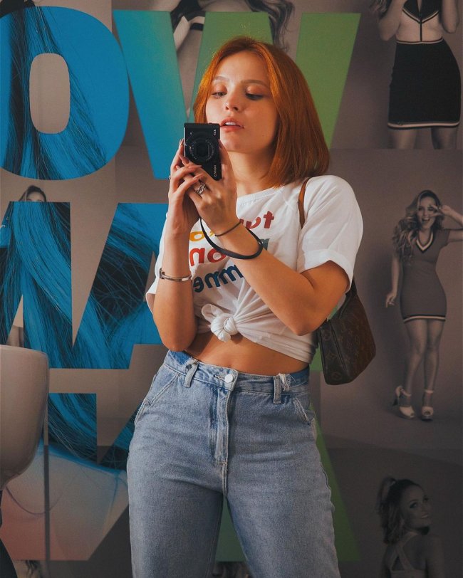 Larissa Manoela frente a espelho fotografando look do dia com camiseta branca, calça jeans e bolsa baguete