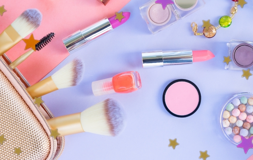 Produtos de maquiagem como batom roxo, laranja, rosa claro, blush, pinceis, necessaire e sombra, espalhados em cima de uma base azul e rosa. Estilo de foto flat lay.