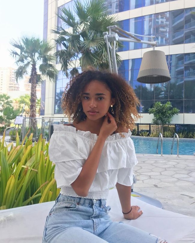 Mélanie Thomas Now United Mélanie, do Now United, com o cabelo solto. Usando camisa branca com babados, calça jeans clara, posando com uma das mãos apoiada no chão e a outra no canto de seu rosto.