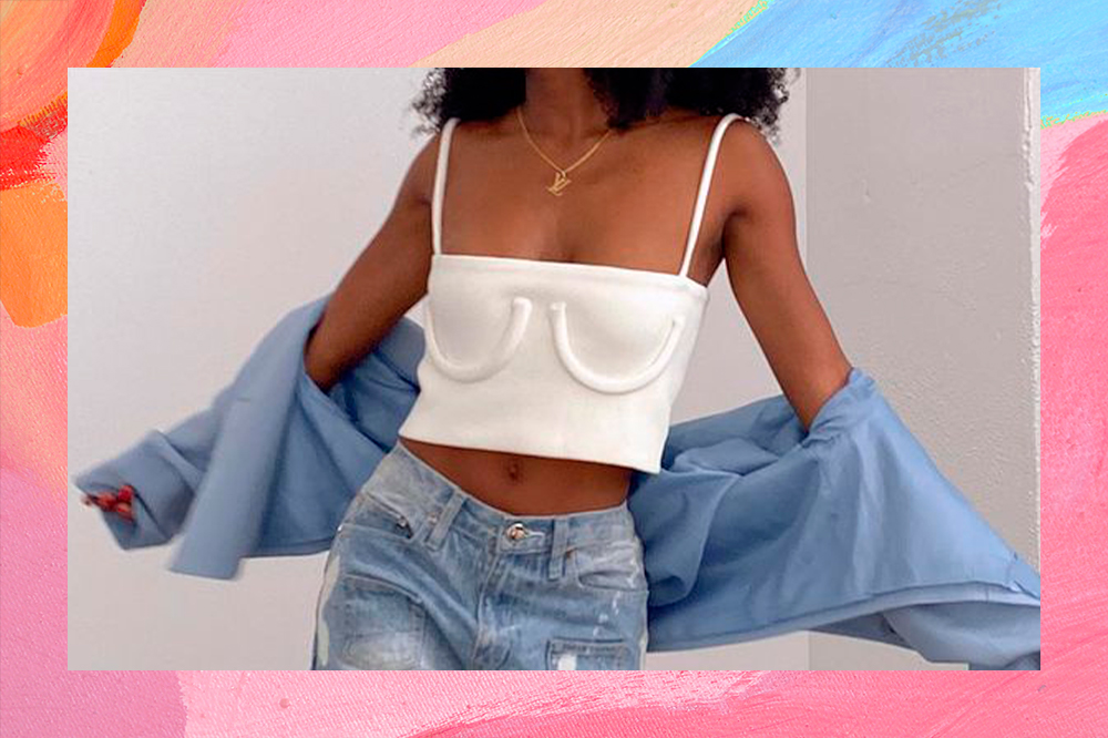 Garota usando top cropped off white com desenho que imita um sutiã com calça jeans de cintura média e uma jaqueta azul caída nos braços. Na foto, dá apenas para ver do pescoço para baixo até a altura do quaril.