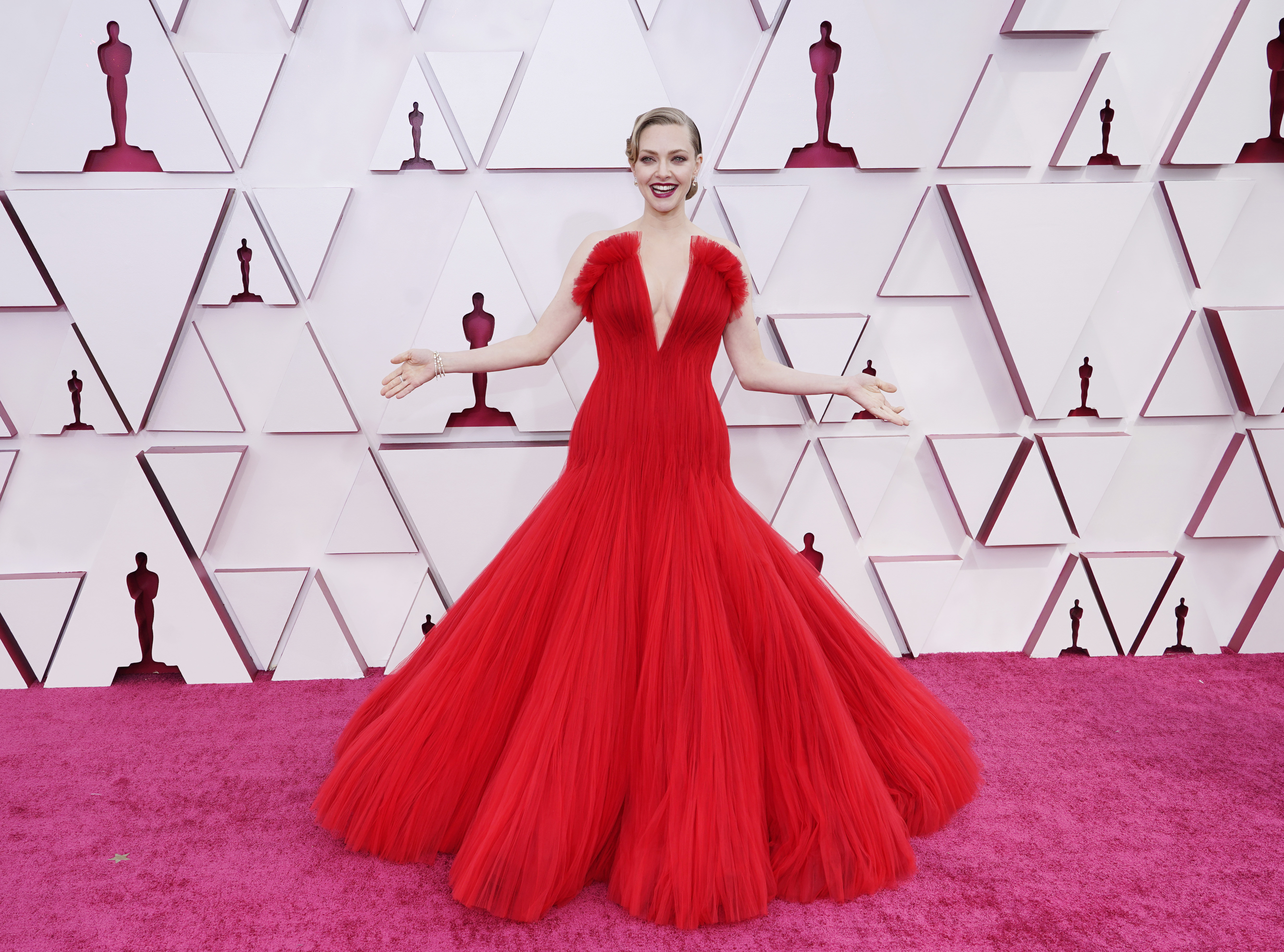 Amanda Seyfried no Oscar 2021. Ela está usando um vestido vermelho de tule com saia volumosa e decote V profundo. Com batom vermelho e cabelo preso em um coque baixo clássico, ela está em pé, com os braços abertos e sorrindo.