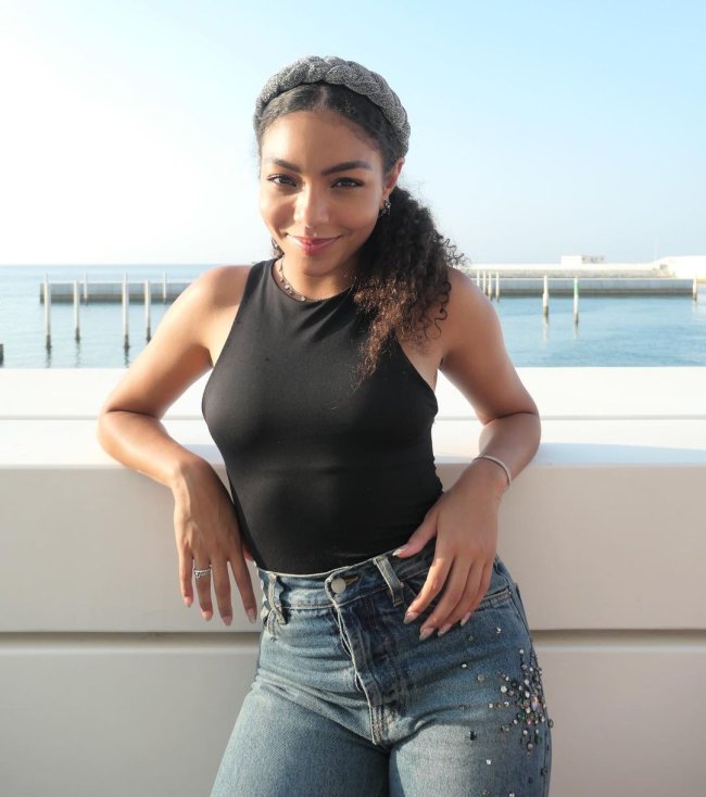 any-gabrielly-rabo-de-cavalo-baixo Cantora e dançarina Any Gabrielly posando apoiada em um muro com os dois braços, usando body preto, calça jeans e tiara em seu cabelo preso em um rabo de cavalo baixo.
