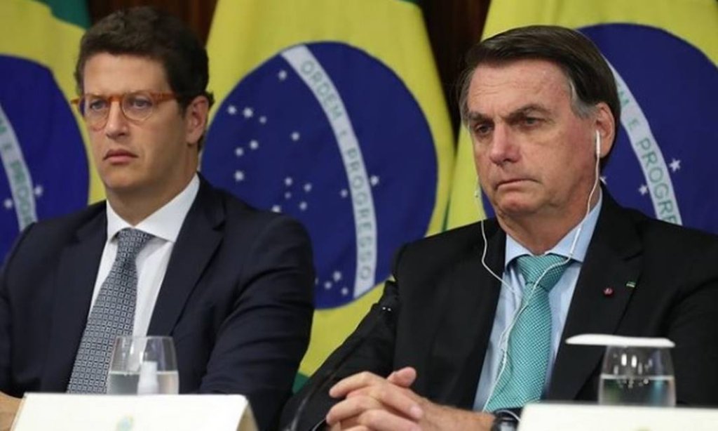 Jair Bolsonaro sentado ao lado de Ricardo Salles durante Cúpula do Meio Ambiente. Ao fundo, aparece a bandeira do Brasil