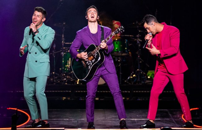 Jonas Brothers se apresentando em Detroid com a turnê Happiness Begins; o Nick veste um conjunto azul turquesa, o Kevin um roxo e o Joe um pink