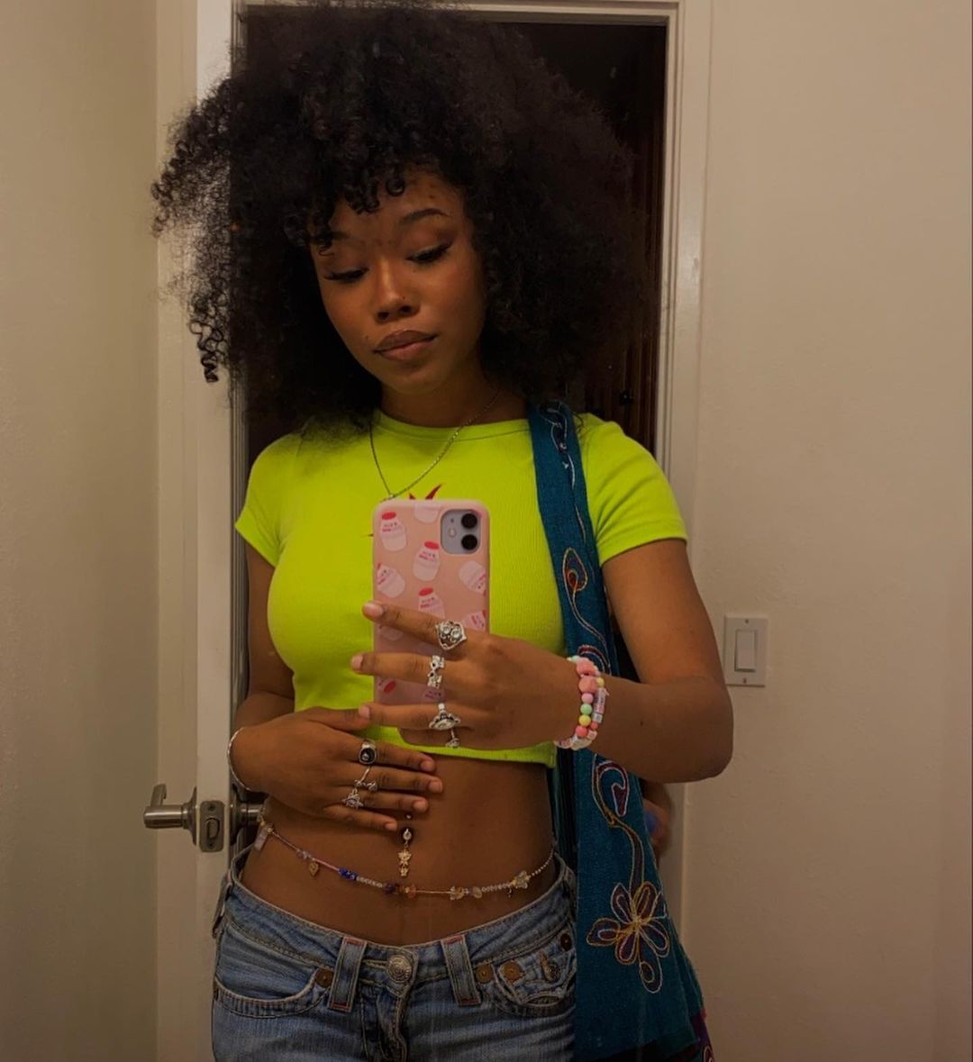 Garota tirando foto com celular em frente ao espelho. Ela usa um top cropped verde neon, calça jeans de cintura baixa, bolsa azul e cinto de miçangas na barriga.