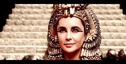 Gif da atriz Elizabeth Taylor como Cleopatra usando adornos dourado ao redor da cabeça delineado grosso com detalhes dourados ao redor dos olhos e piscando para a câmera.