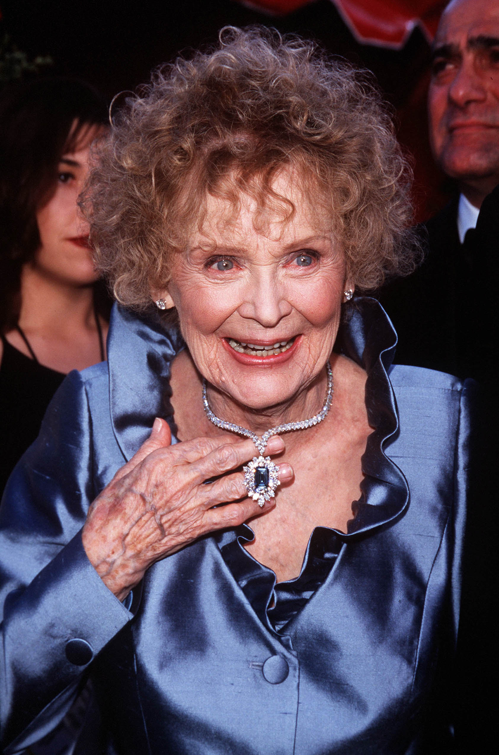 gloria-stuart-oscar-1998 A atriz Gloria Stuart no Oscar de 1998. Ela está usando uma camisa azul de seda com babados na gola. Uma das suas mãos está segurando seu colar, que é todo de diamantes e tem um pingente com uma pedra azul no meio. Ela está sorrindo e olhando levemente para o lado direito.