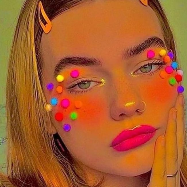 Jovem posando para foto com close em seu rosto, com maquiagem com o canto do olha iluminado e muito blush nas bochechas com bolinhas coloridas coladas ao redor dos olhos, batom rosa neon e uma das mãos apoiadas no rosto.