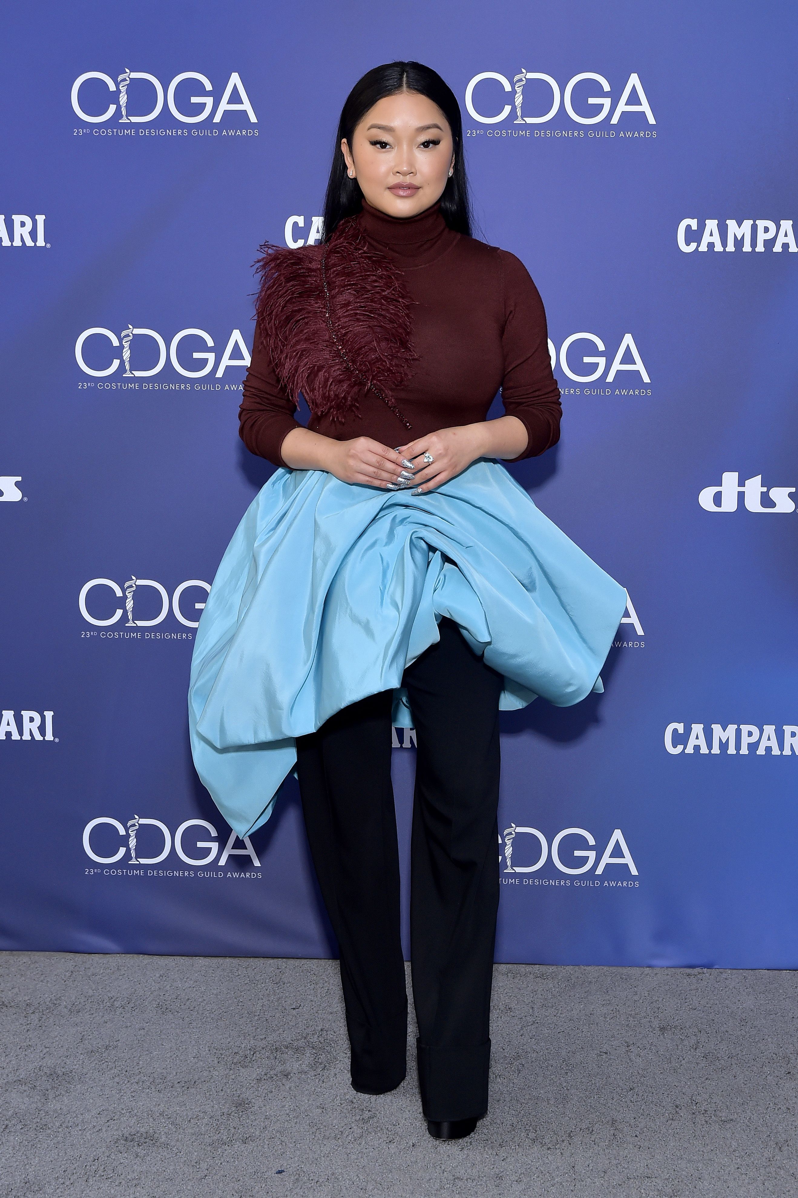 Lana Condor usando suéter bordô de gola alta que tem uma aplicação de plumas em um dos ombros, uma calça pantalona azul-marinho e, por cima, uma saia azul turquesa volumosa e assimétrica. Ela está olhando para frente, com a expressão séria, os braços dobrados e as mãos unidas na frente do corpo.