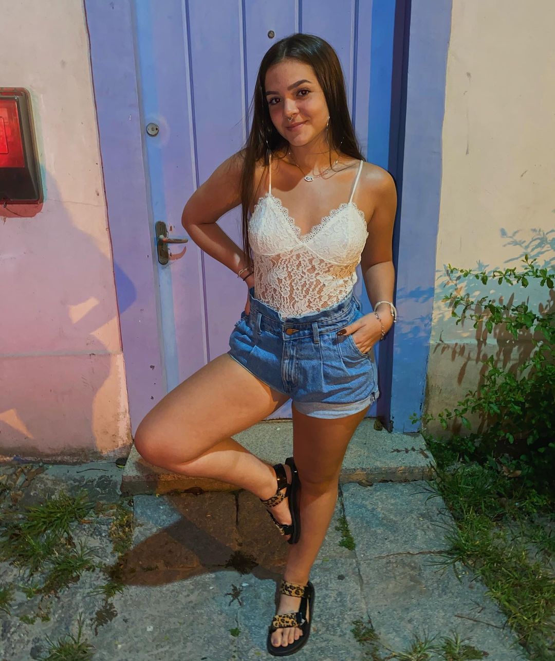 Mel Maia usando look com body de renda branco, short jeans modelo clochard e uma papete preta com estampa de oncinha nos pés. Ela posa em pé, com uma das pernas dobradas e apoiada na outra, formando um quatro, uma das mãos no bolso, e a outra na cintura. Ela está sorrindo, sem mostrar os dentes.