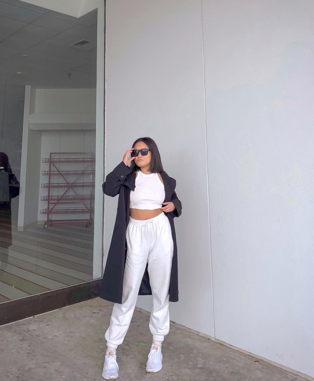 Garota usando top cropped branco com calça de moletom e tênis brancos. Ela está com um sobretudo preto por cima do look, e uma das mãos segura os óculos escuros, enquanto a outra está na cintura.