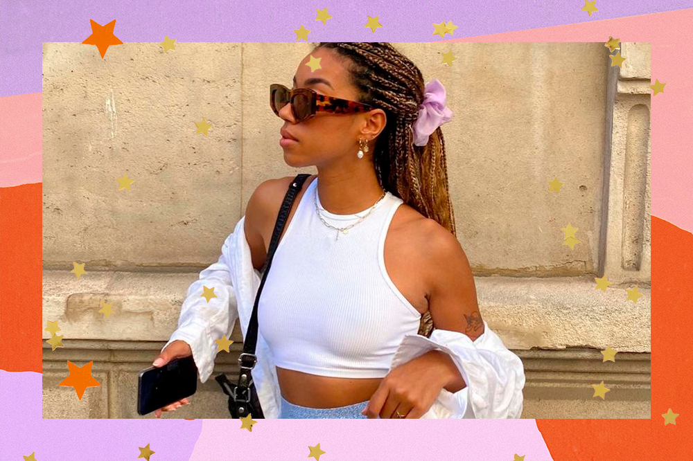 Foto de garota usando top cropped branco com camisa branca por cima. Ela está olhando para o lado, com expressão facial séria, usando óculos escuros de tartaruga e cabelo preso em um rabo com scrunchie lilás. Há uma bolsa preta pendurada em um dos ombros. Com uma das mãos, ela segura um celular, e a outra está apoiada na altura da cintura. A foto é uma montagem com um fundo colorido em laranja, rosa e lilás, com estrelinhas amarelas.