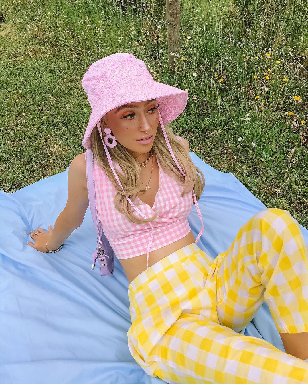 Garota usando look com top cropped xadrez vichy rosa e calça amarela na mesma estampa. Ela está sentada, com os dois braços para trás, uma bolsa lilás pendurada em um dos ombros, além de um chapéu rosa floral e um brinco rosa.