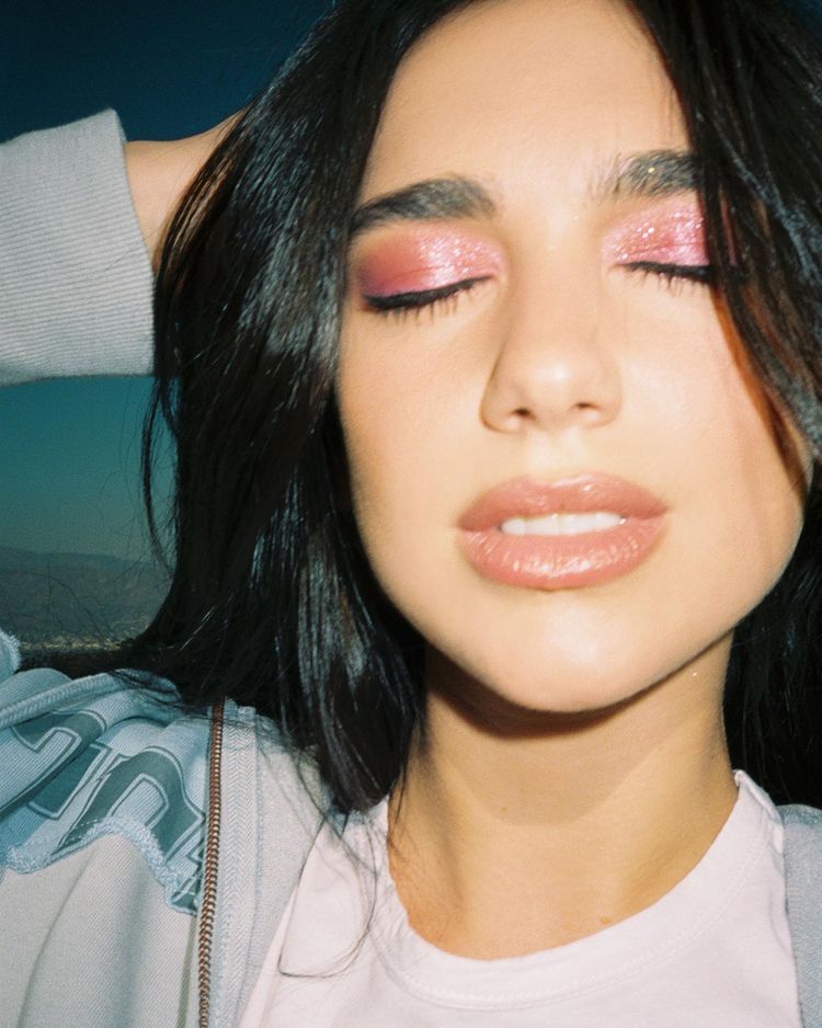 maquiagem-dua-lipa-02 Dua Lipa usando maquiagem com sombra rosa de brilhos e gloss marrom. Ela está com os olhos fechados, boca levemente aberta e uma das mãos na cabeça.