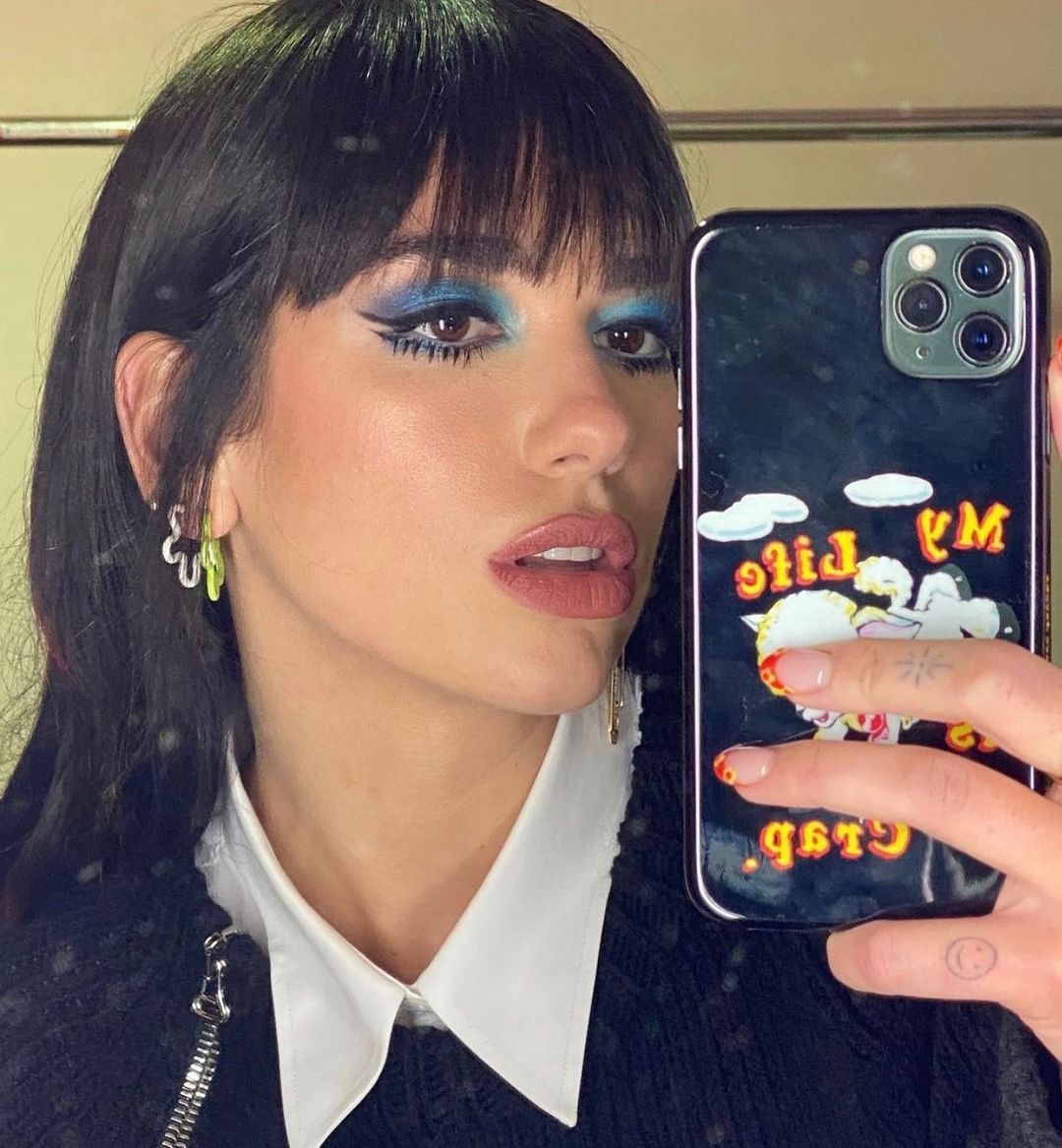 Dua Lipa usando maquiagem com sobra azul e delineado preto, no estilo anos 60. Ela está em frente a um espelho, tirando a foto com o celular próximo ao rosto. Sua expressão facial é séria e a boca está levemente aberta.