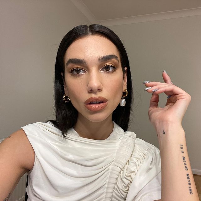maquiagem-dua-lipa-05 Dua Lipa usando maquiagem com sombra marrom esfumada. Ela está com uma blusa branca de um ombro só, uma das mãos levantadas e expressão facial séria com a boca levemente aberta.