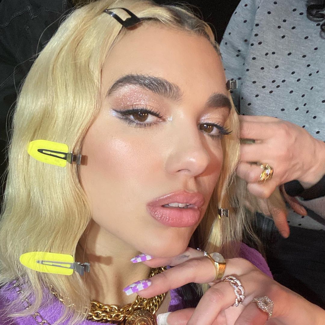 maquiagem-dua-lipa-07 Dua Lipa usando maquiagem com sombra de brilhos e delineado fino. Ela está usando presilhas ao longo do cabelo. Está com expressão facial séria, olhando levemente para cima e uma das mãos no queixo.