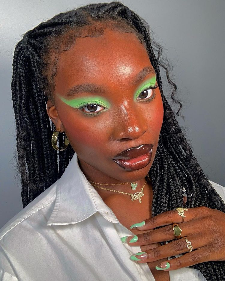 Garota usando maquiagem com sombra verde, uma camisa branca e mix de colares dourados. Sua mão, com as unhas com nail art verde, está apoiada perto do pescoço. Sua expressão facial é séria.