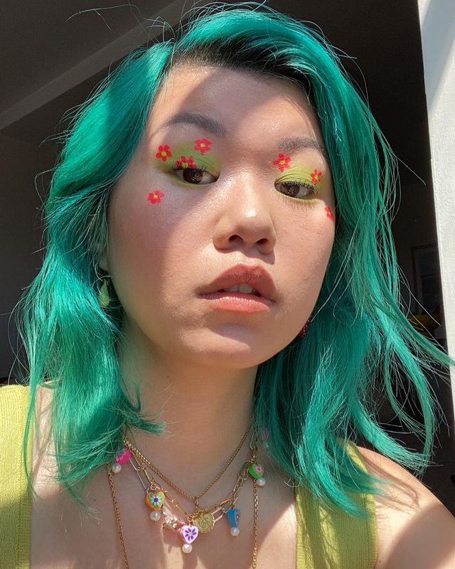 Garota usando maquiagem com sombra verde e florzinhas laranjas. Seu cabelo e sua regata são verdes e ela usa mix de colares. Ela está olhando para baixo, com expressão facial séria e boca levemente aberta.