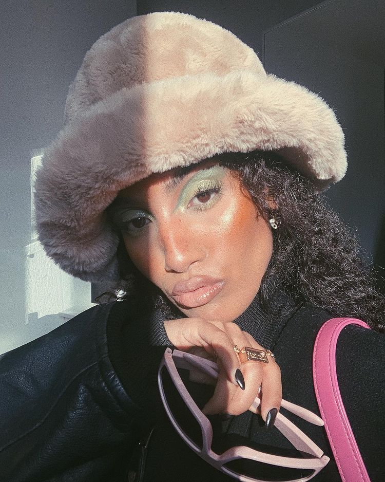 Garota usando maquiagem com sombra verde, chapéu de pelúcia e casaco preto. Ela está fazendo um bico com a boca e, com uma das mãos, segura um óculos de sol cor-de-rosa.